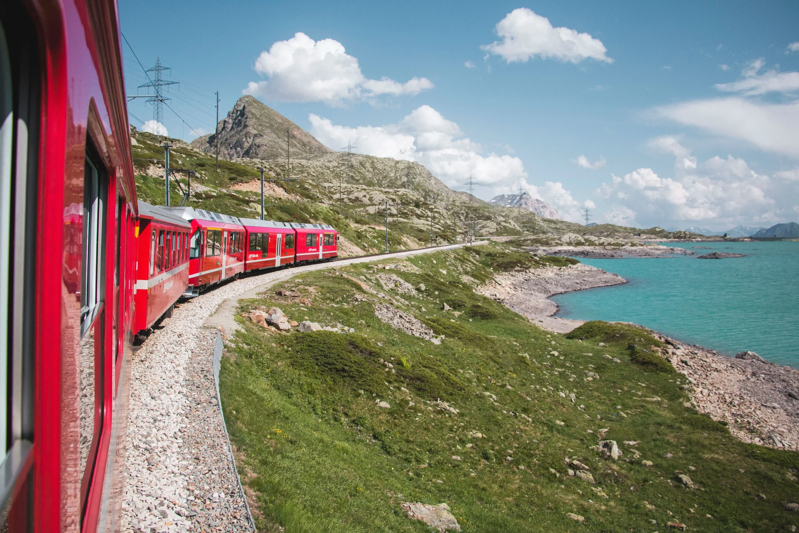 Bernina Express railway journey: Red train traversing railway with mountain lake beside, framed by lush green mountains in the backdrop.