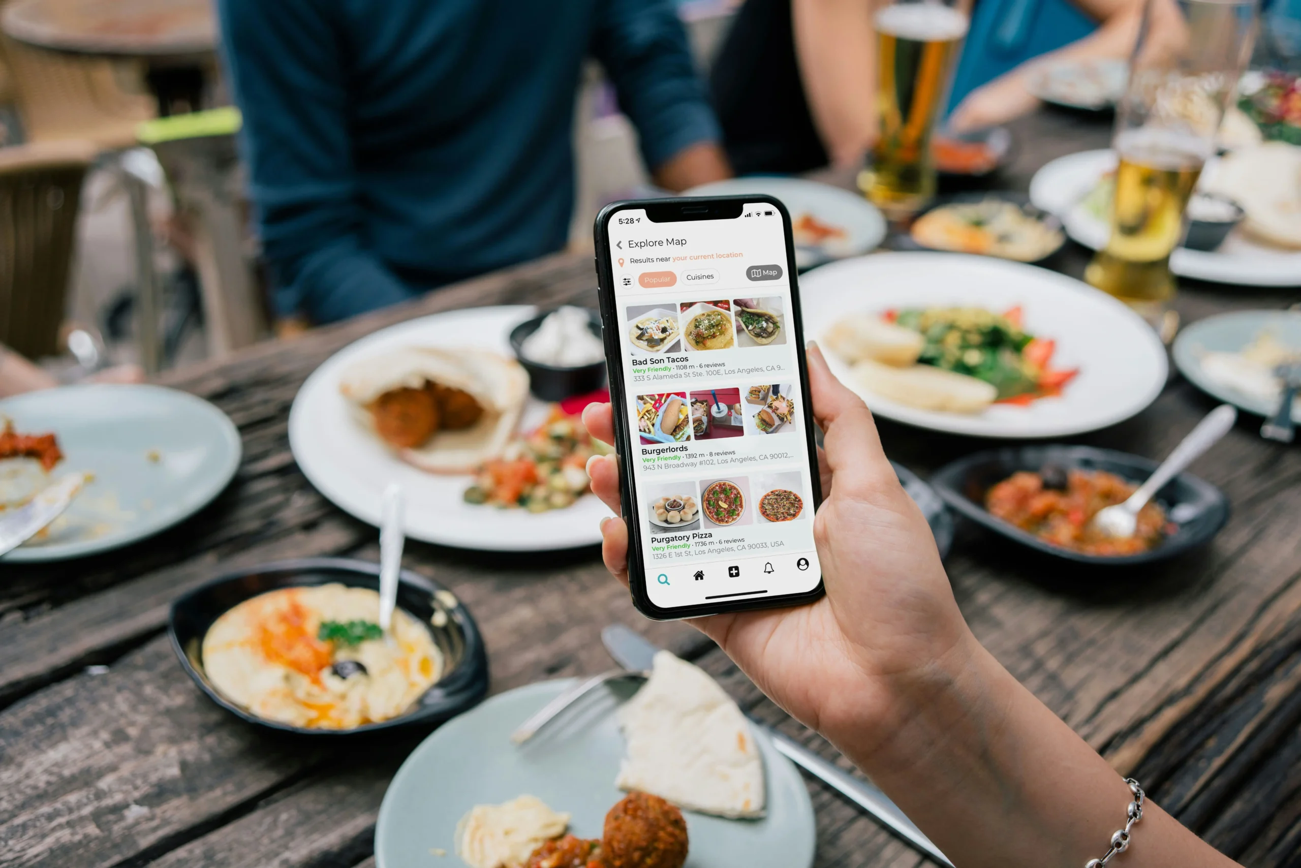 A person is using an app to find the best restaurant in the area. In the background, there is a table with food.